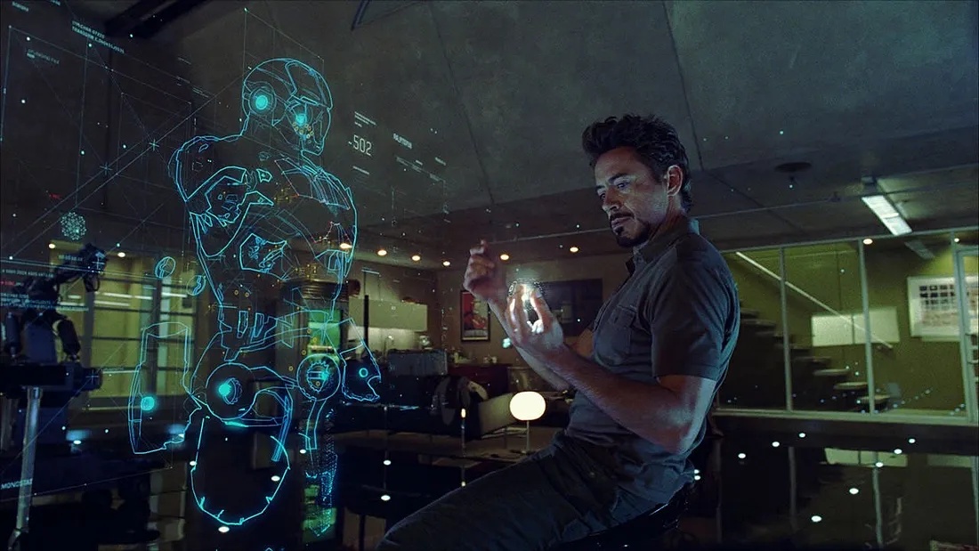 Man in a dark shirt sits in a modern office, manipulating a glowing blue holographic armored-suit projection.