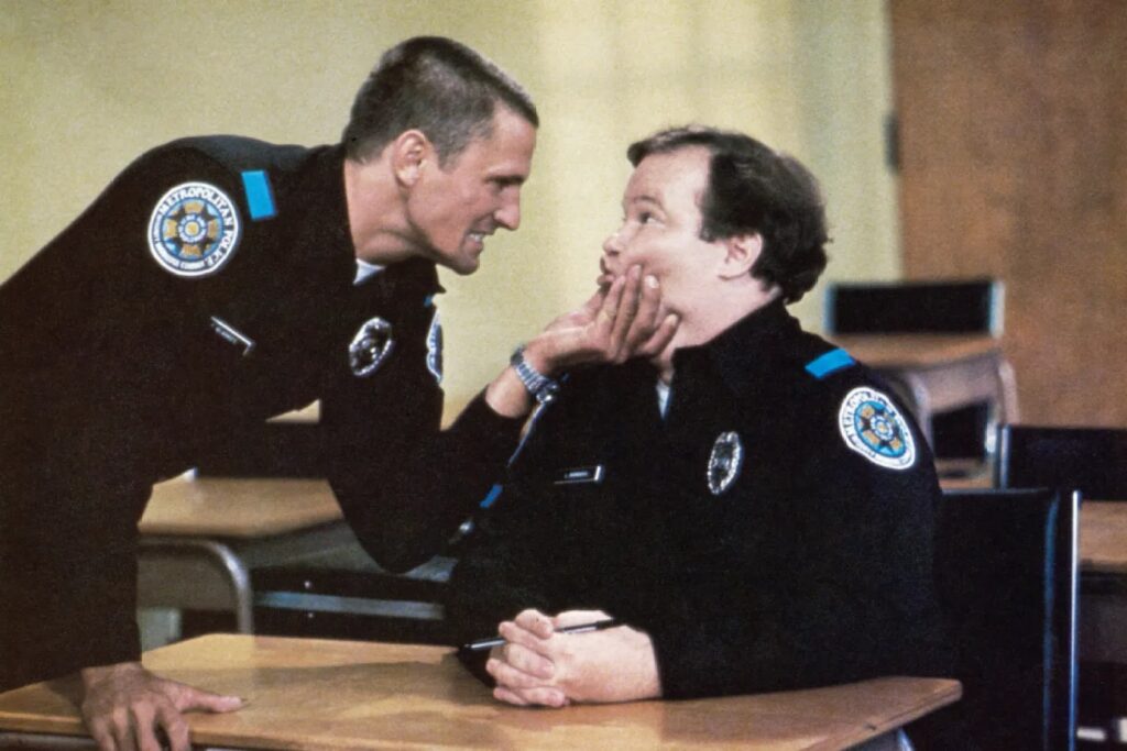 Two police officers in uniform lean toward each other at a desk; one cupping the other’s face as they talk in a classroom setting.