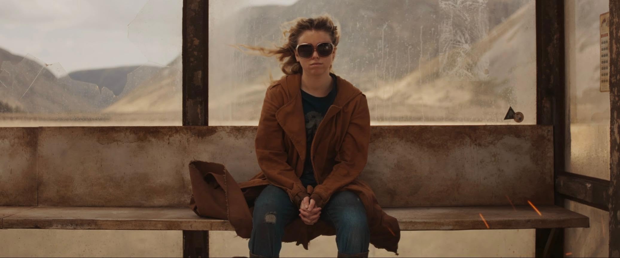 Woman in a brown coat and sunglasses sits on a weathered bus-stop bench, wind-swept hair and mountains behind her.