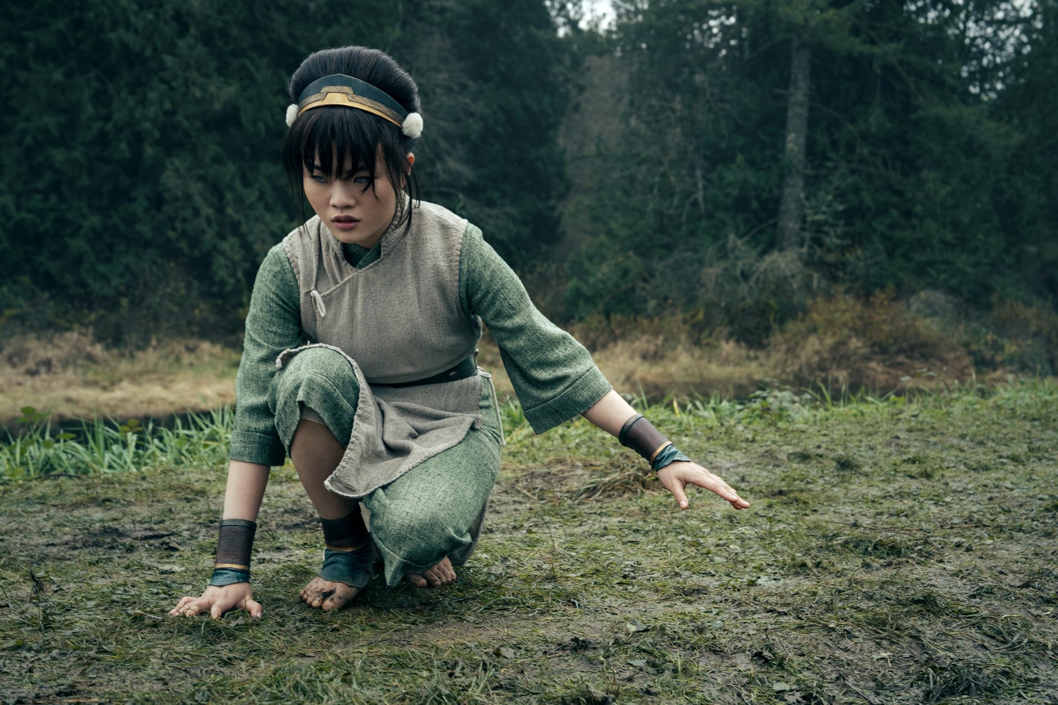 Person in a green tunic and headband crouches on muddy ground in a forest clearing, looking downward.