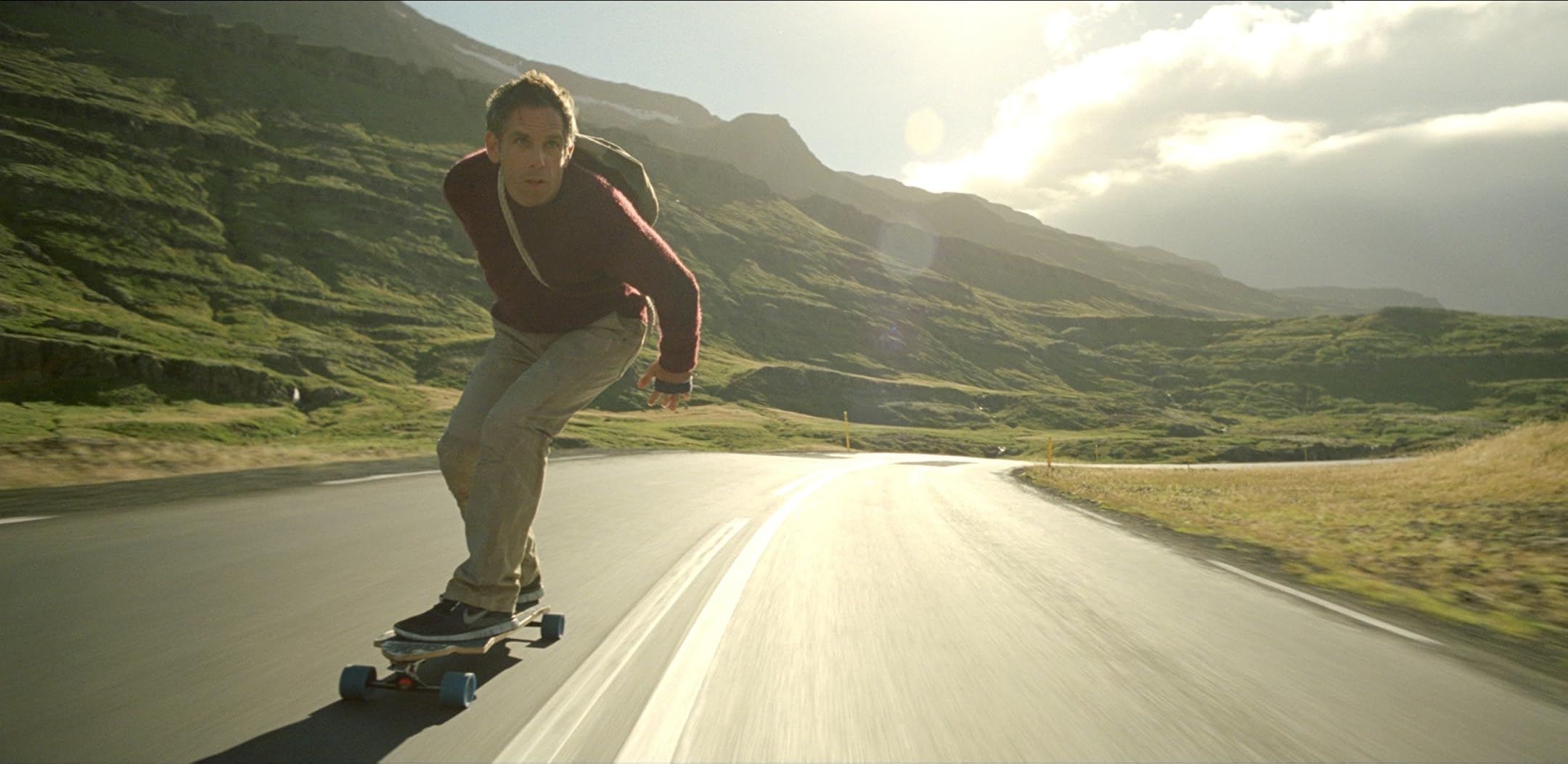 Skateboarder in a burgundy hoodie leaning into a turn on a winding mountain road with green hills.