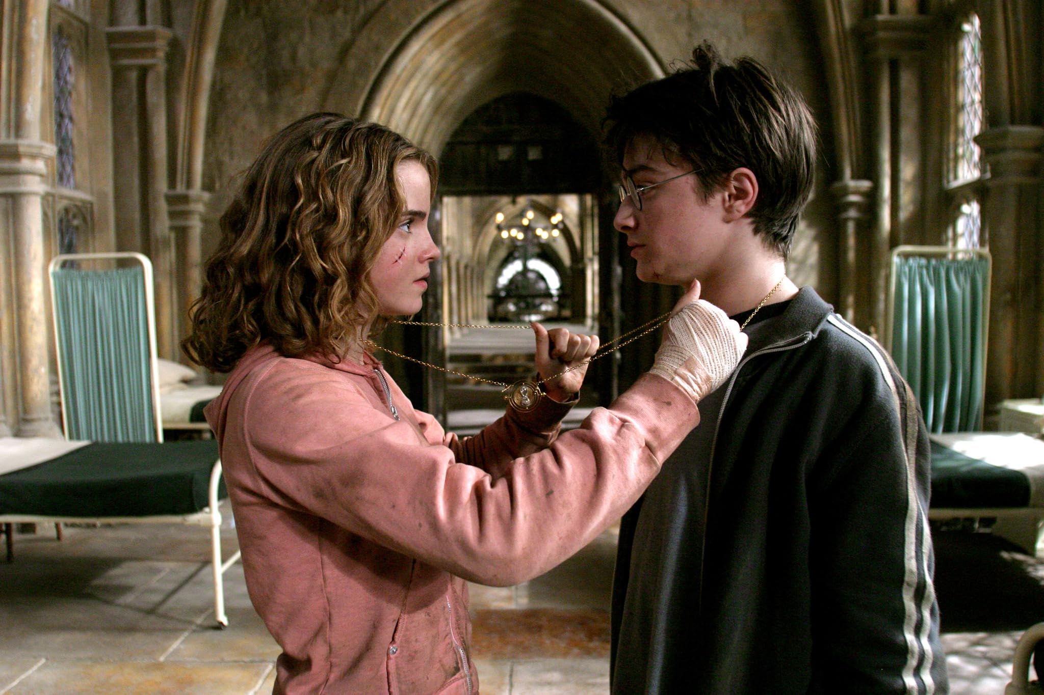 Two young people stand face to face in a cathedral-like hallway, holding a gold chain between them as if tying or presenting it.