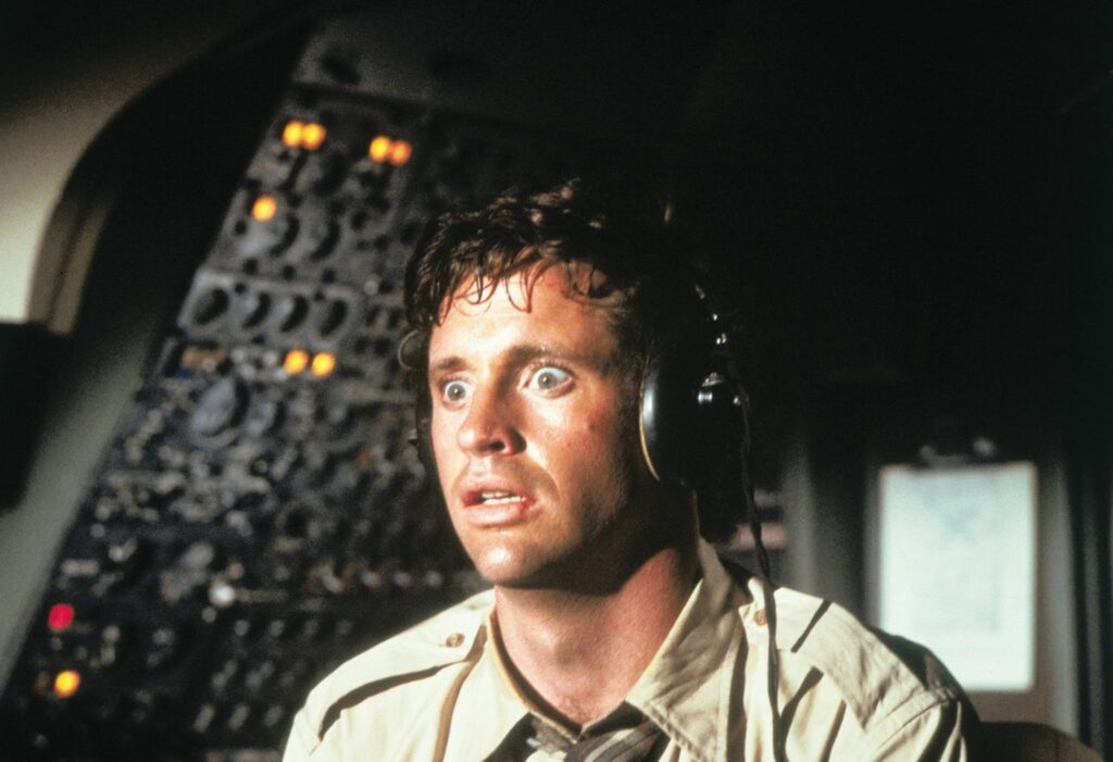 Pilot in cockpit wearing a headset, looking alarmed with illuminated controls behind him.