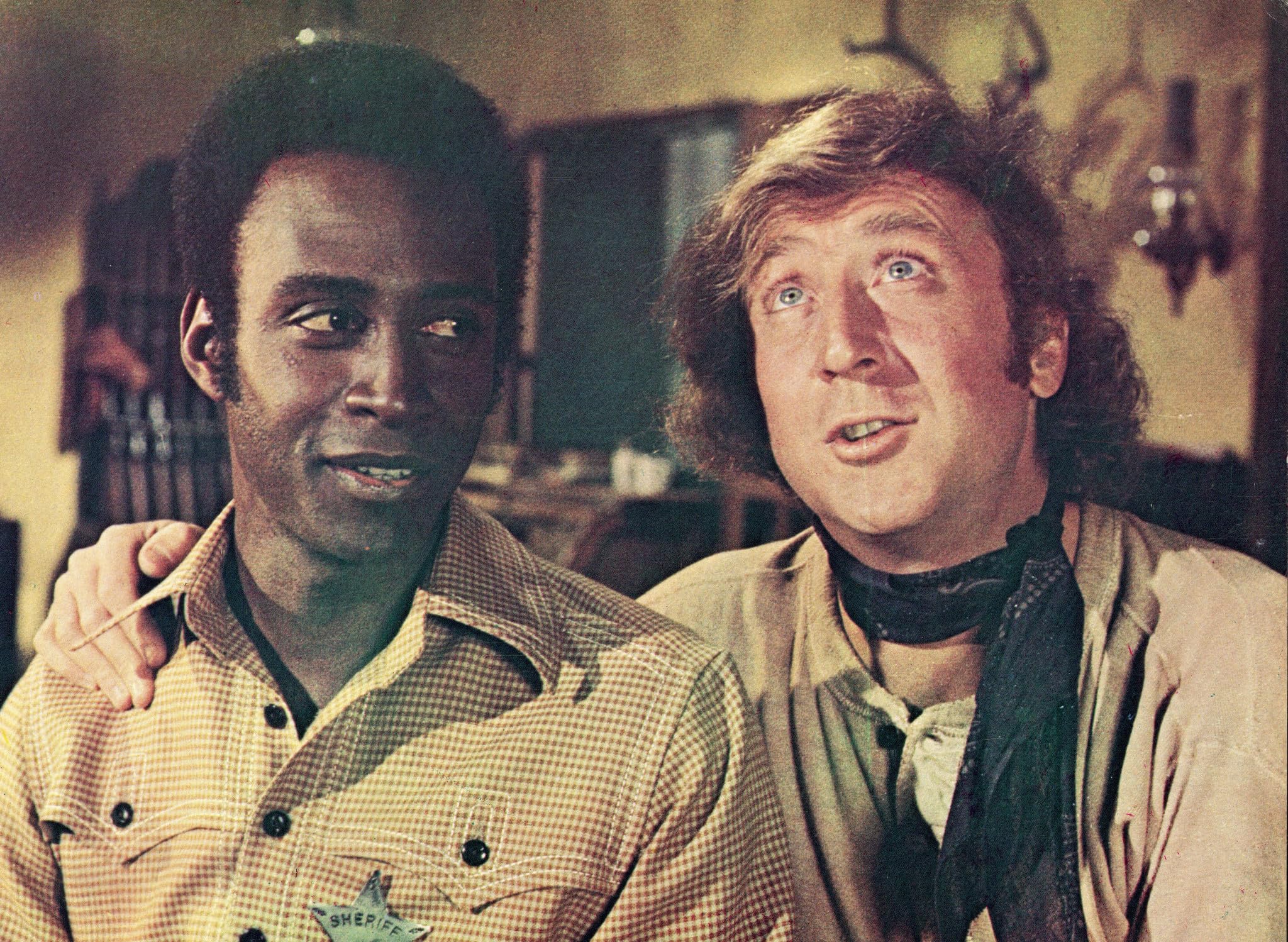 Two men in plaid shirts stand close, one with his arm around the other, looking up and smiling indoors.