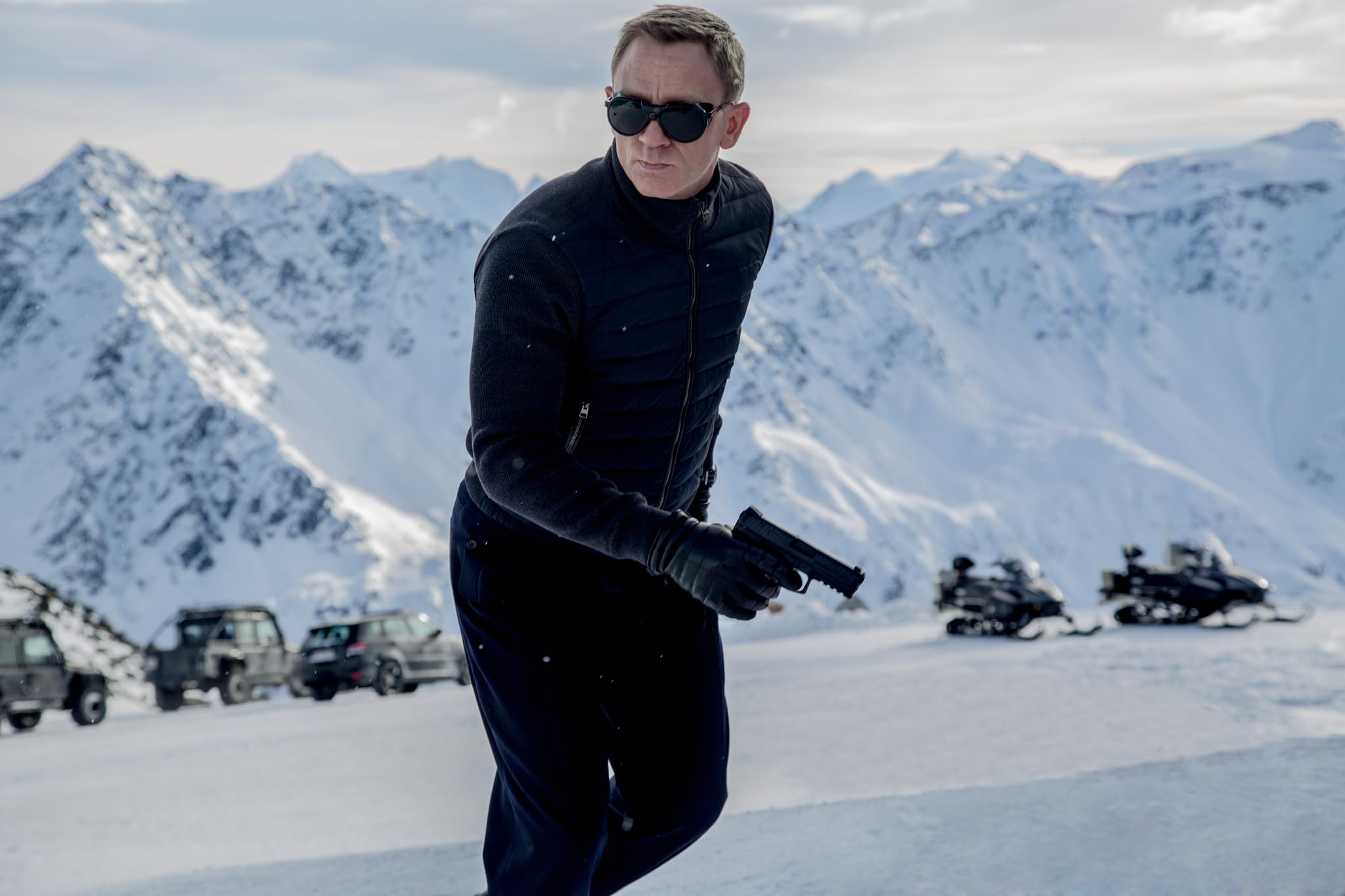 Man in a black vest and gloves stands on a snowy mountain, looking to the side with sunglasses on. Behind him are parked cars and snow-covered peaks.