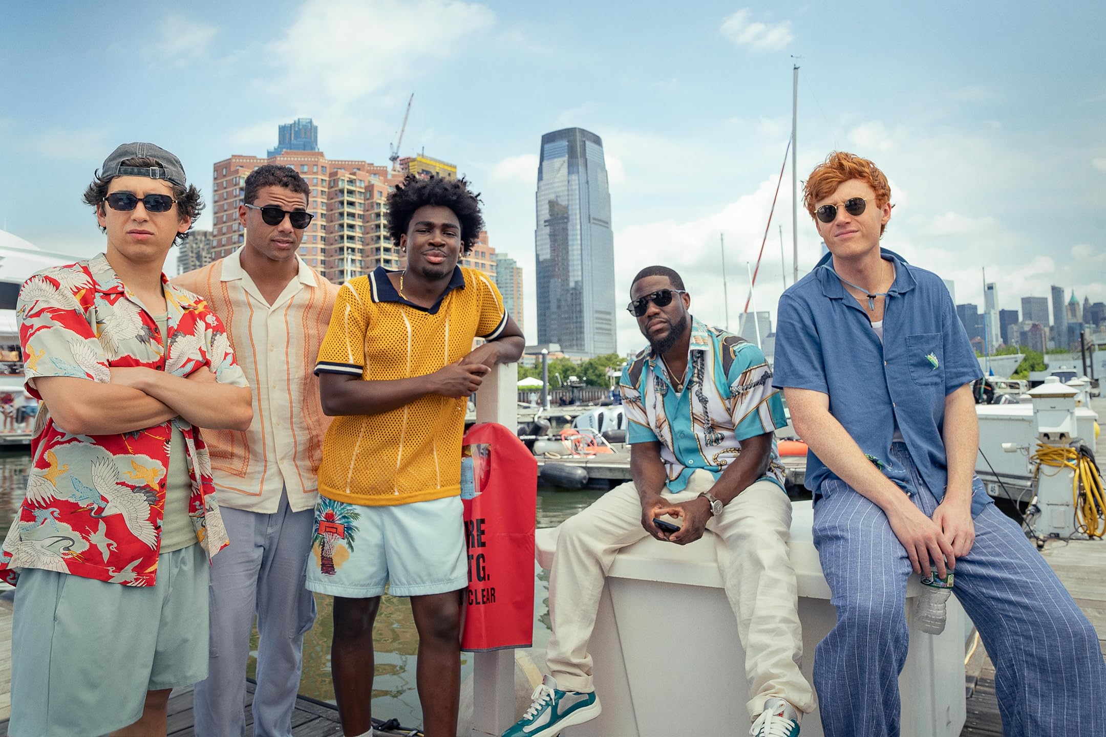 Five young men in colorful shirts and sunglasses pose on a marina dock with a city skyline in the background.