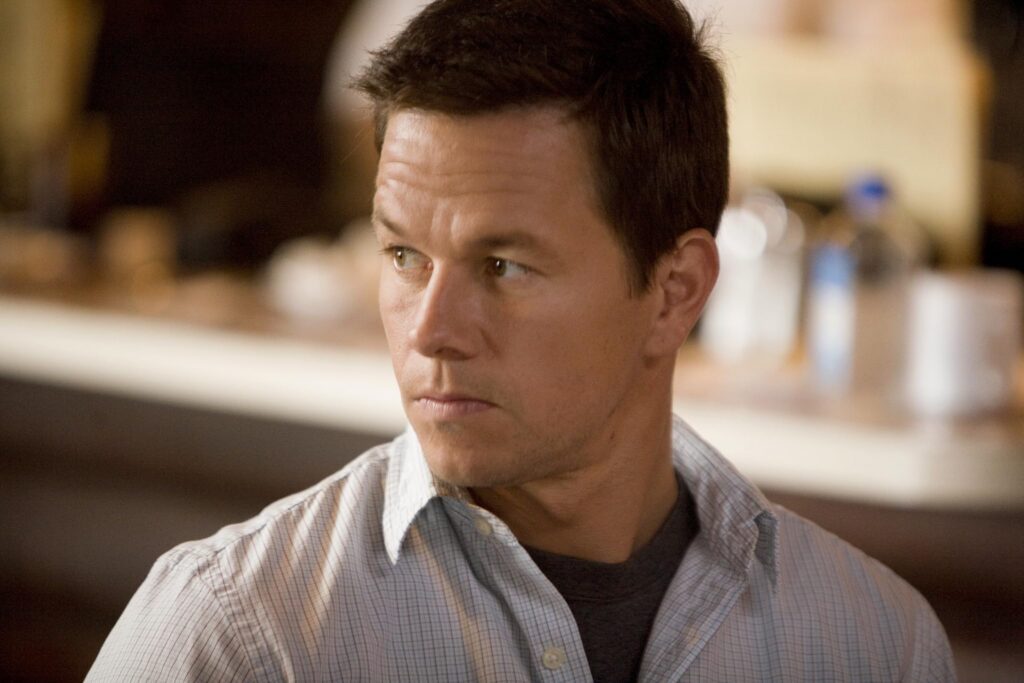 Man with short dark hair in a light checkered shirt, looking to the left with a neutral expression in an indoor setting that's softly blurred in the background.