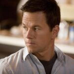 Man with short dark hair in a light checkered shirt, looking to the left with a neutral expression in an indoor setting that's softly blurred in the background.