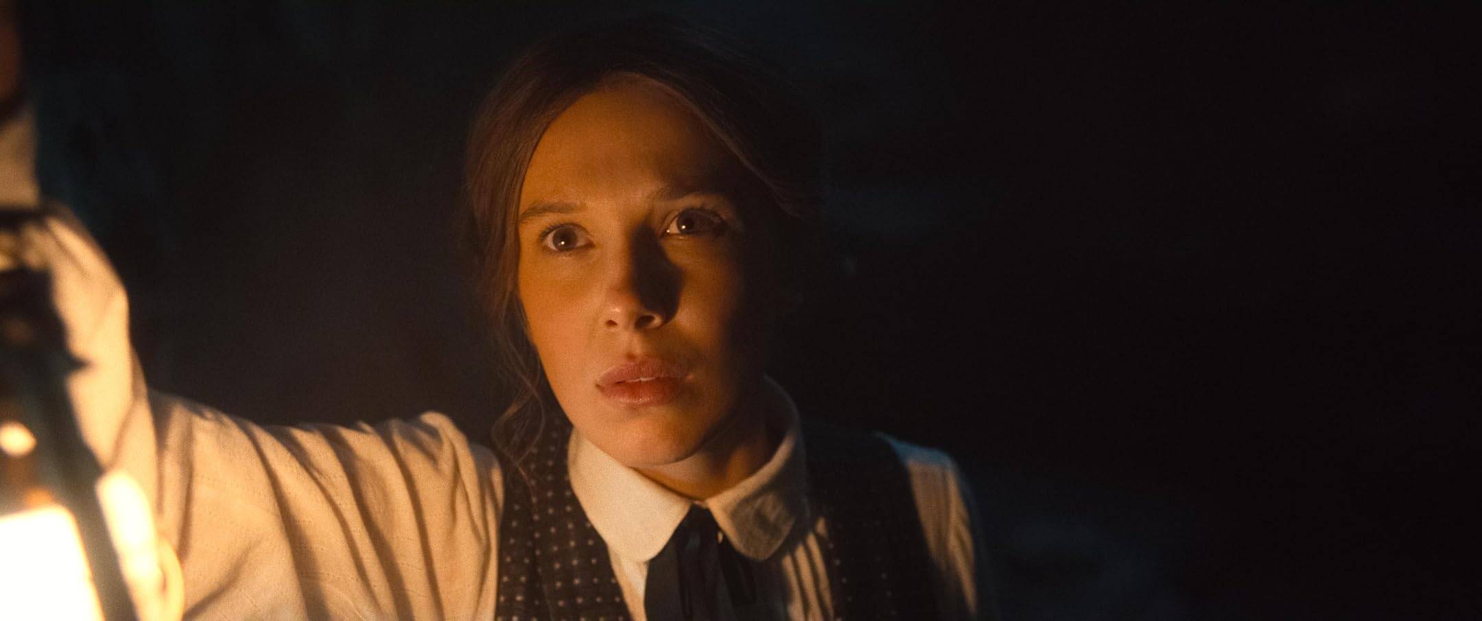 Woman in a white blouse with a dotted vest, lit by a lantern, looks toward the light with a serious expression in a dark setting.