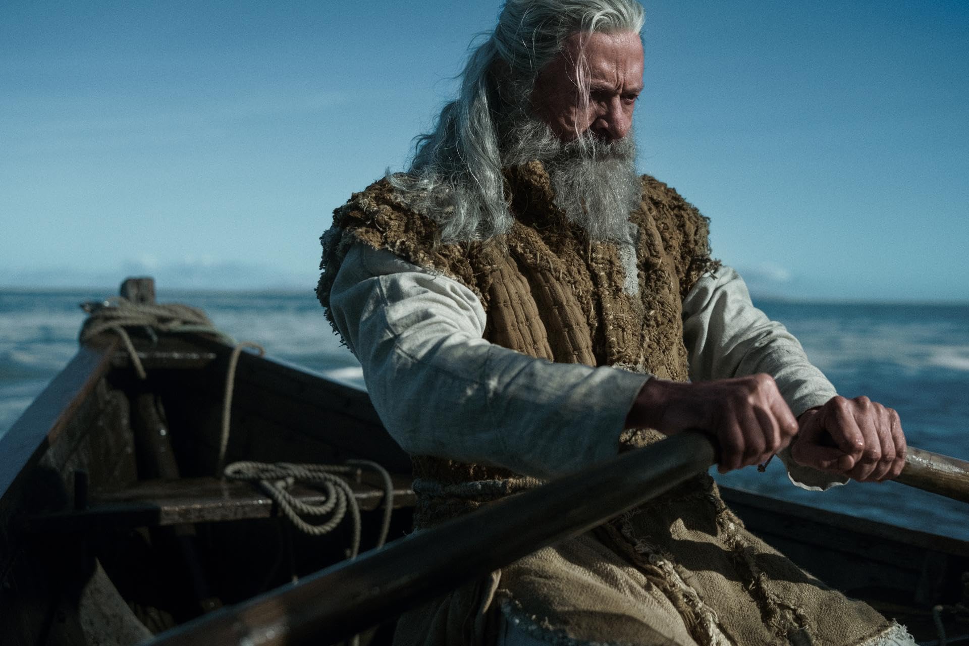 Older man with long gray hair and beard sitting in a boat, gripping the oars against a blue sea backdrop.