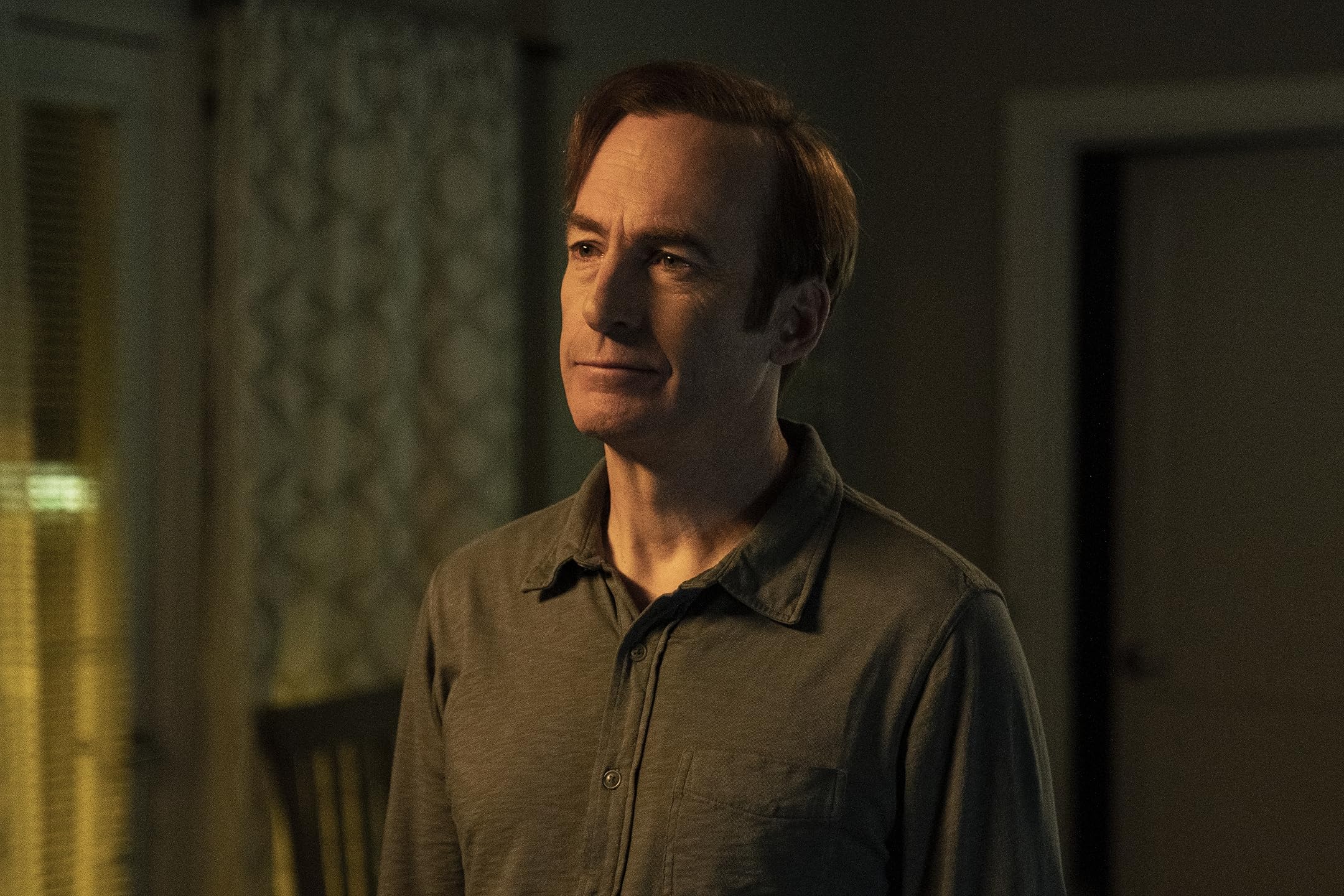 Man in a casual button-up shirt stands in a dimly lit room, looking slightly to the right with a calm expression.