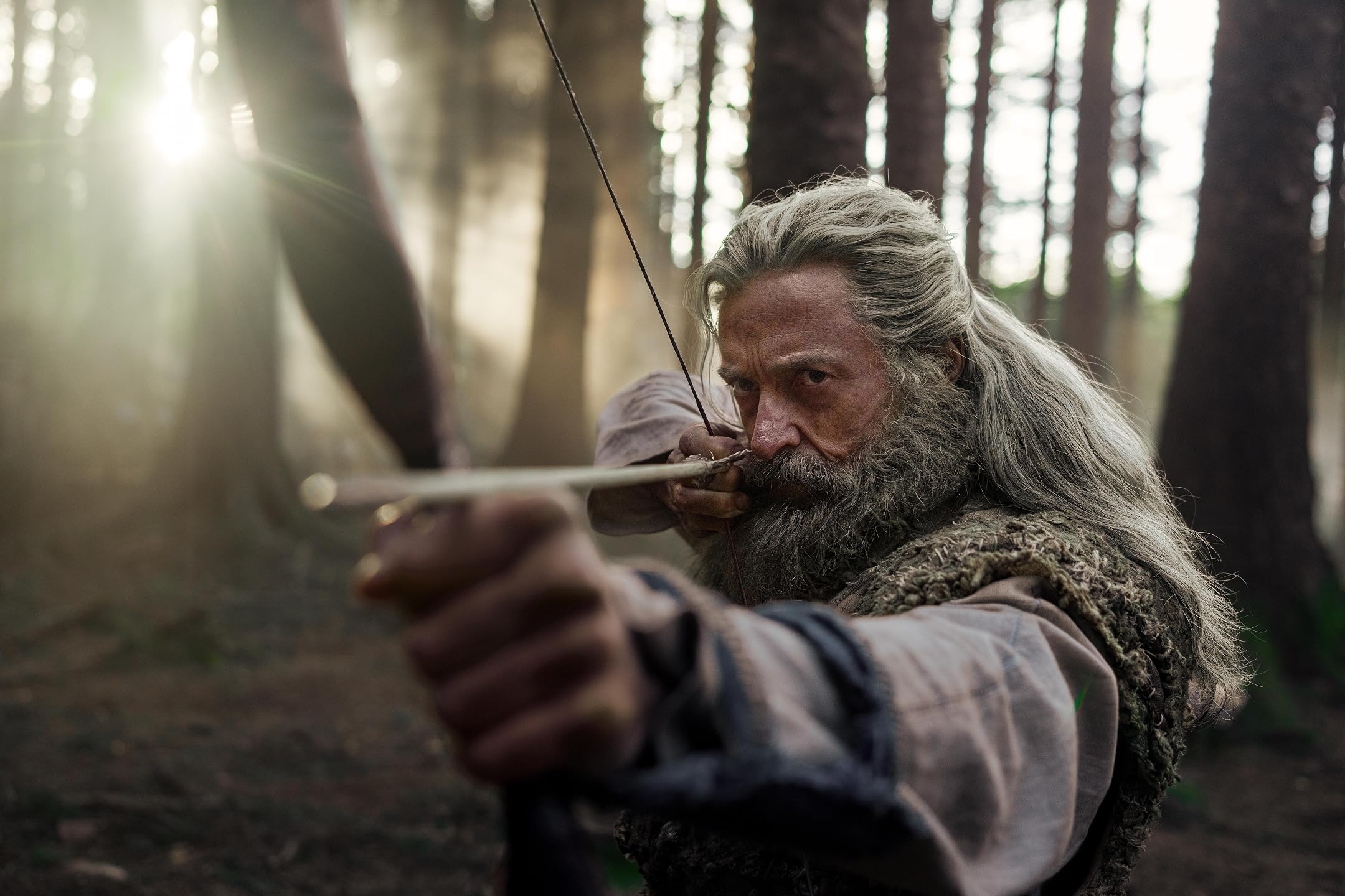 Older man with long gray hair draws a bow in a sunlit forest, focusing on his target.