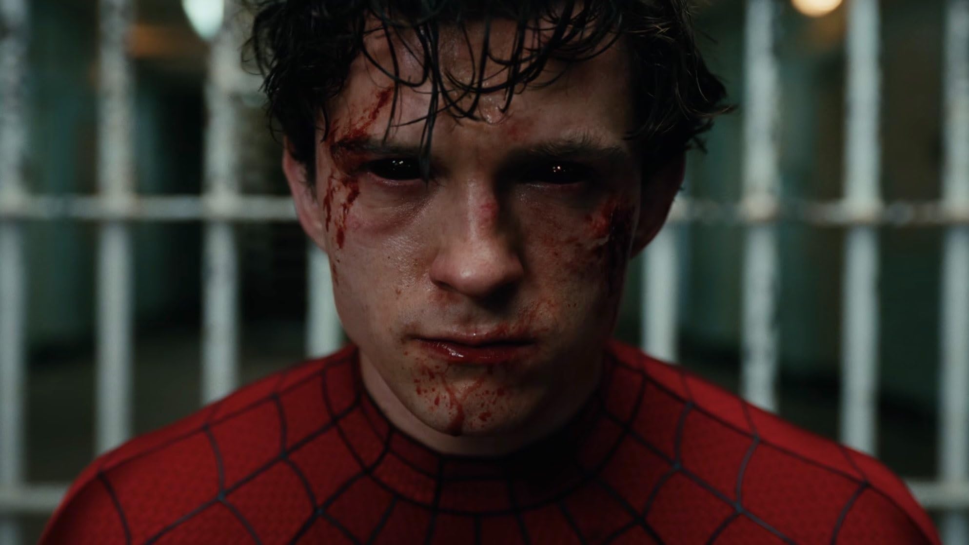 Close-up of a young man in a red Spider-Man suit with blood around his eyes and mouth, standing behind metal bars.