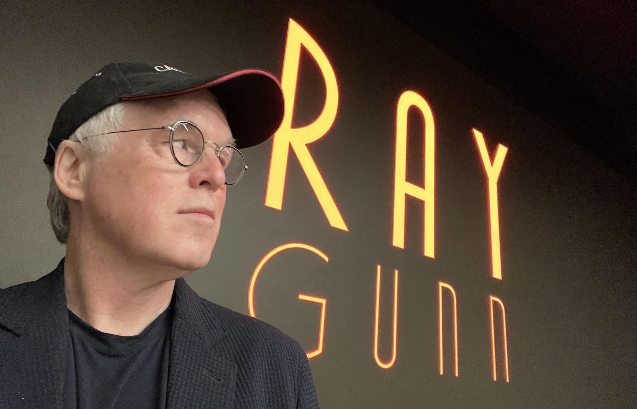 Man in a black cap and glasses looks at a vertical orange neon sign to his right.
