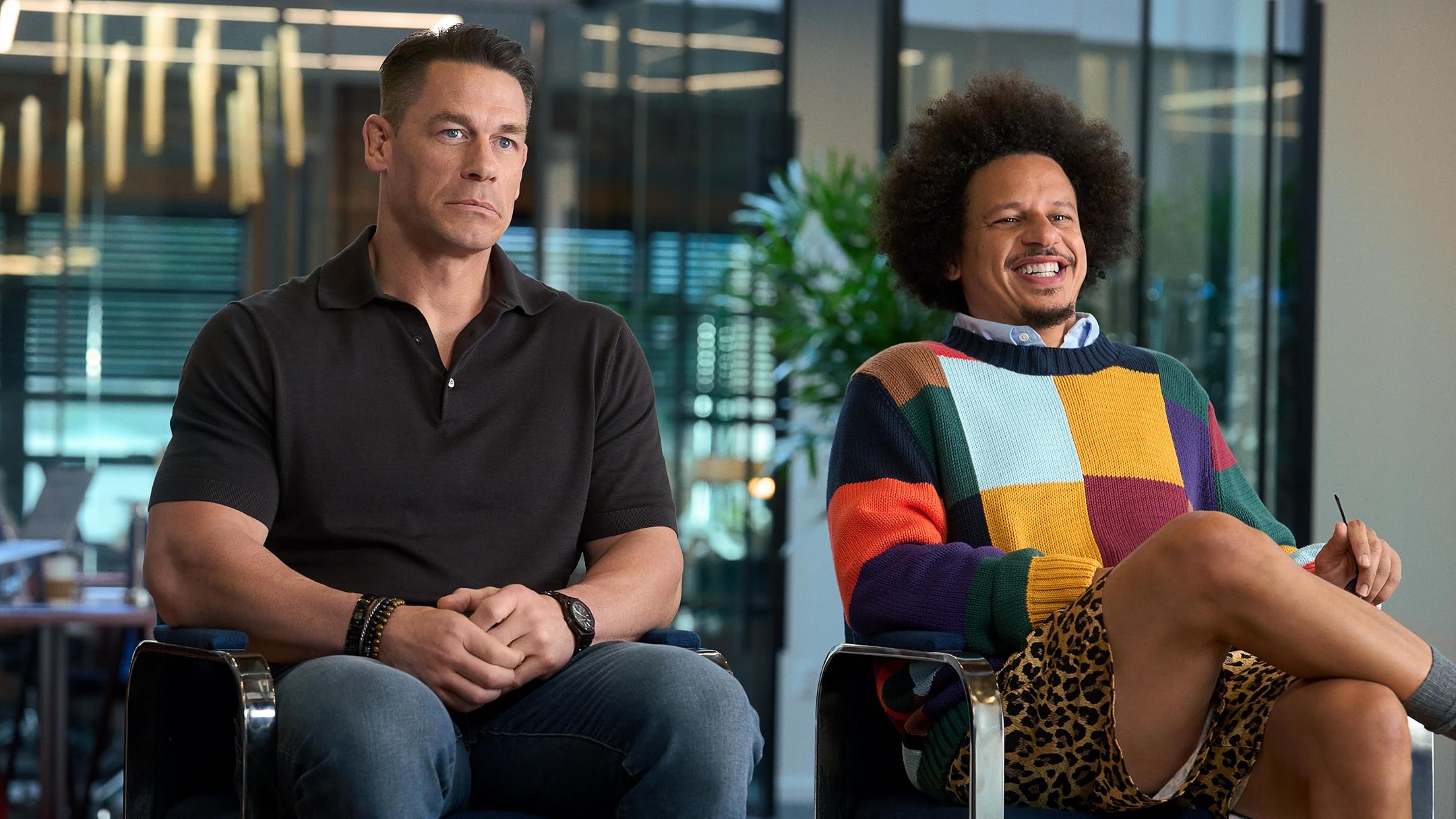 Two men sit side by side in an office, one serious in a black polo and the other smiling in a colorful knit sweater, engaged in conversation.