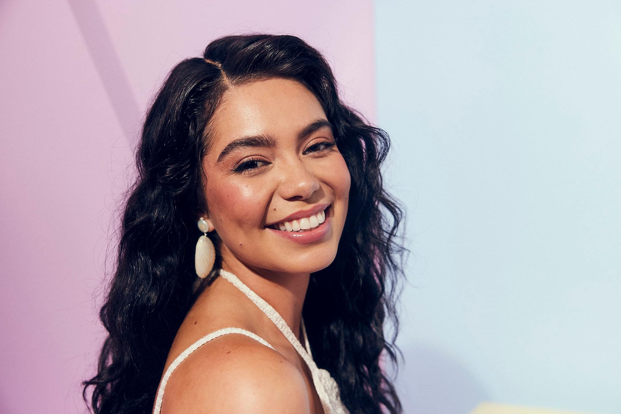 Smiling woman with long dark hair against a pastel pink and blue backdrop, wearing a white strap top.
