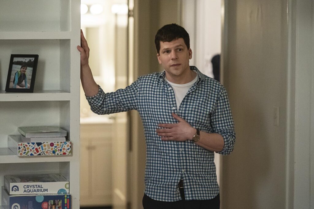 Man in a blue plaid shirt stands in a doorway, left hand resting on a shelf with a framed photo and toy boxes nearby.