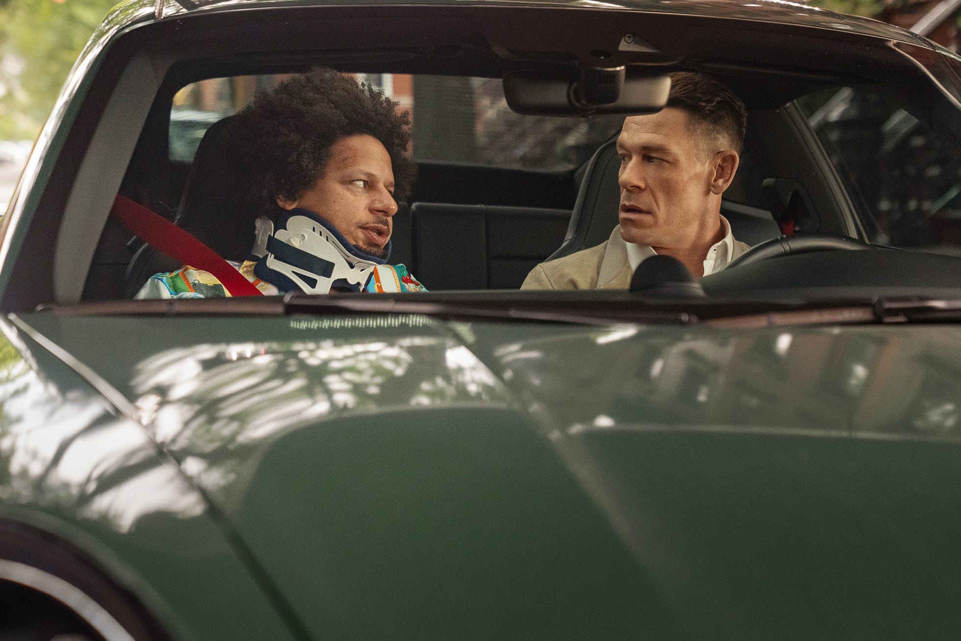 Two men sit in a car’s front seats, the passenger wearing a neck brace and talking to the driver across the console.