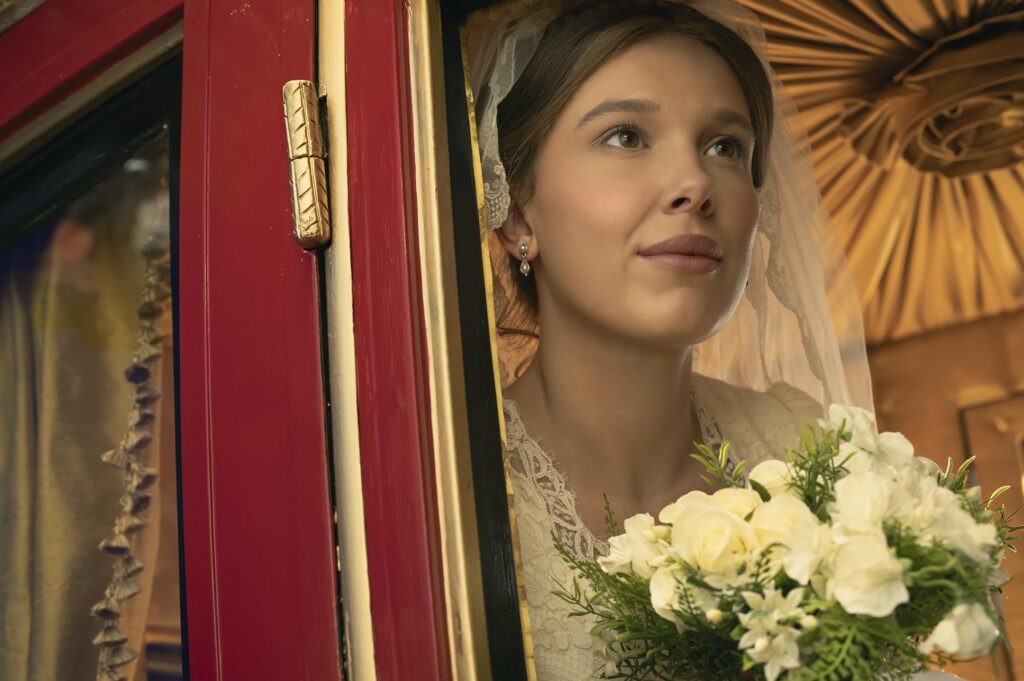 Bride mannequin in a wedding dress peeking through a red doorway, holding a bouquet of white roses and greenery in a shop window display.