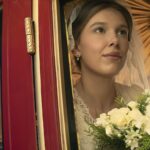 Bride mannequin in a wedding dress peeking through a red doorway, holding a bouquet of white roses and greenery in a shop window display.