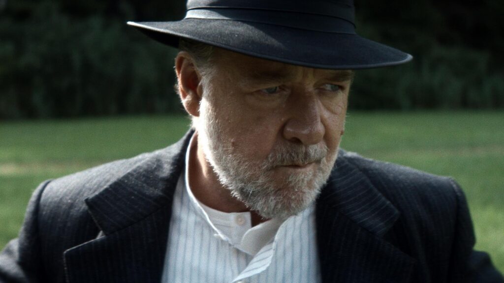 Close-up of an older man with a gray beard wearing a dark hat and pinstripe blazer, outdoors in a grassy area, looking to the side.