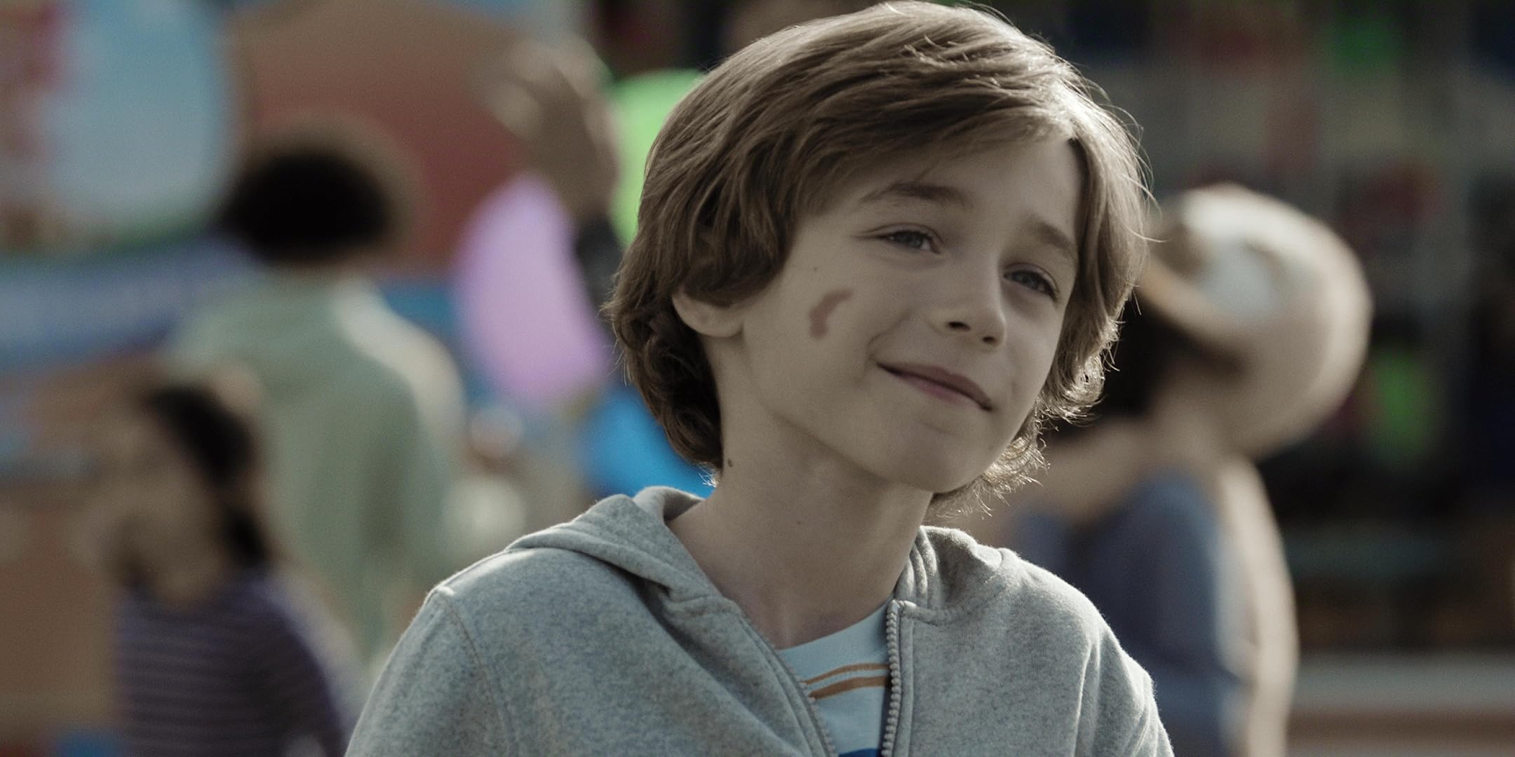 Boy with a small face paint mark on his cheek, smiling slightly, wearing a gray hoodie at an outdoor event with blurred people in the background
