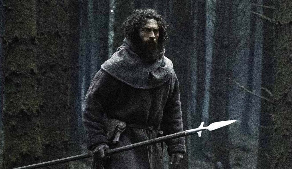 Man in a heavy gray cloak with a hood, beard, standing in a dark forest, gripping a long spear.
