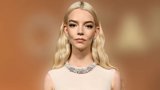 Portrait of a pale-dressed blonde woman wearing a diamond necklace, standing on a warm orange backdrop (runway style).