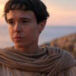 Person with short dark hair wrapped in a light brown shawl stands along a rocky coastline at sunset, looking contemplative.