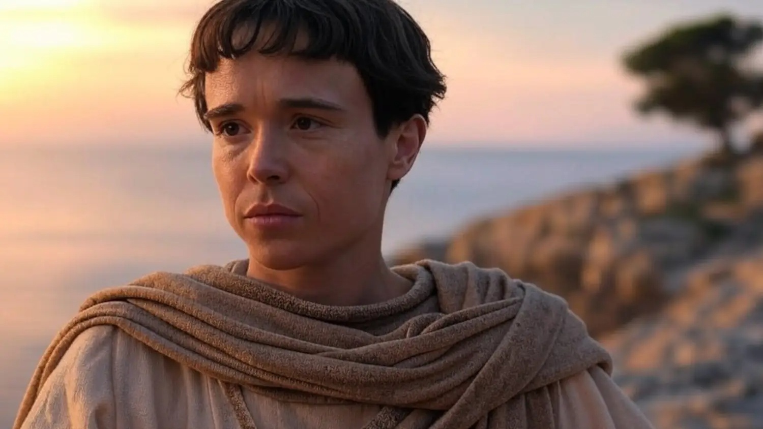 Person with short dark hair wrapped in a light brown shawl stands along a rocky coastline at sunset, looking contemplative.