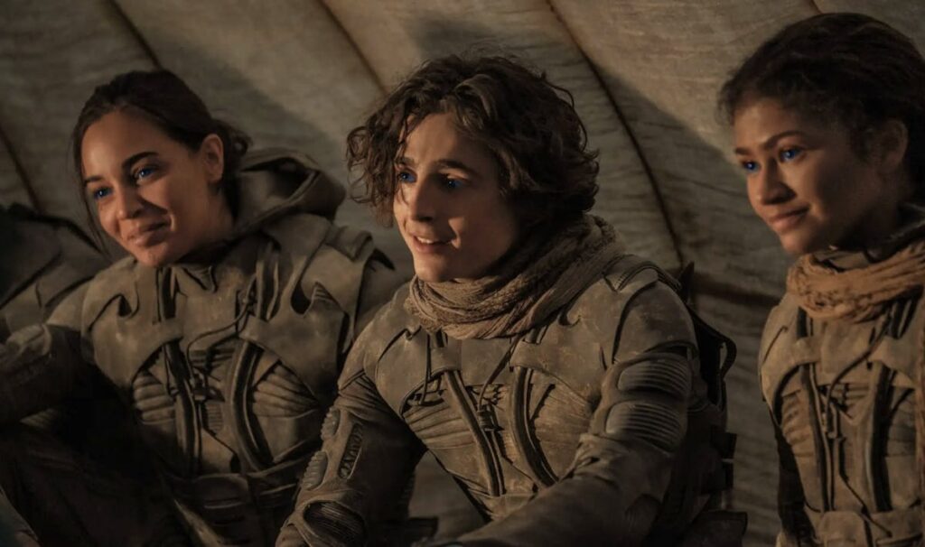 Three astronauts in dusty beige suits sit inside a curved shelter, looking ahead with calm expressions.
