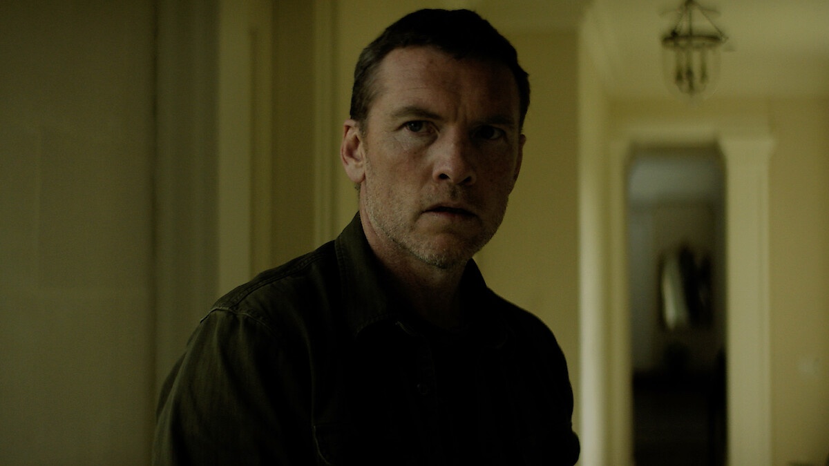 Close-up of a man in a dark shirt looking worried, standing in a dim hallway, facing the camera slightly off-center.