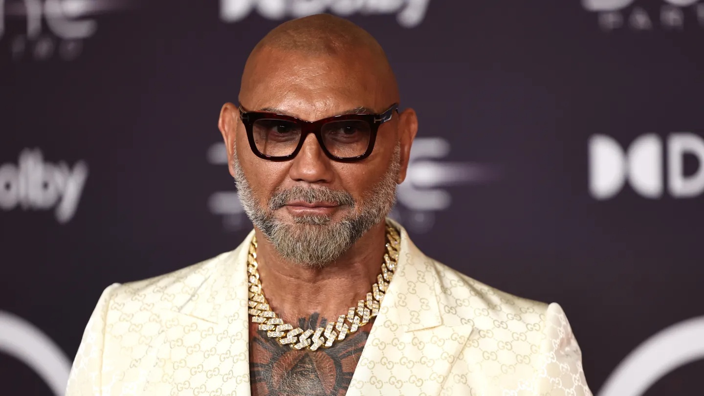 Man with shaved head and gray beard wearing a cream Gucci-pattern suit and a thick gold chain on a red-carpet event backdrop.