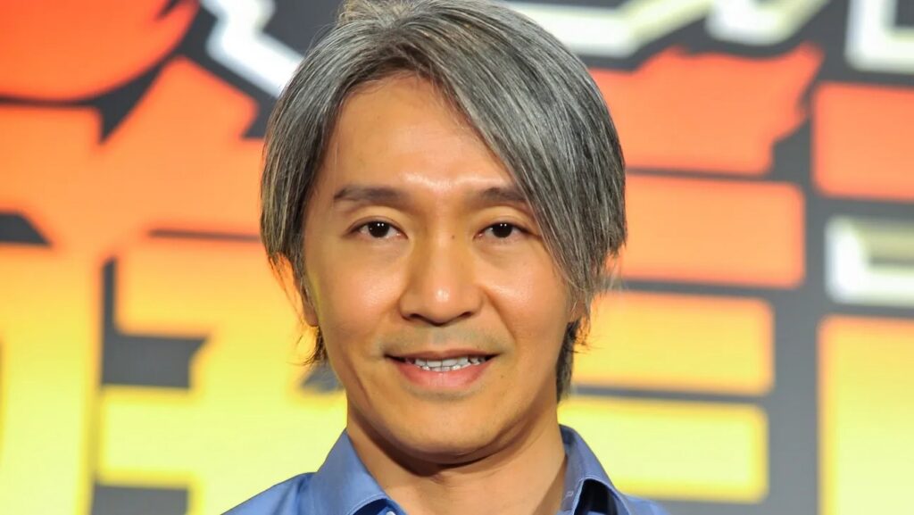 Smiling man with salt‑and‑pepper hair in a blue shirt against an orange‑yellow backdrop. l