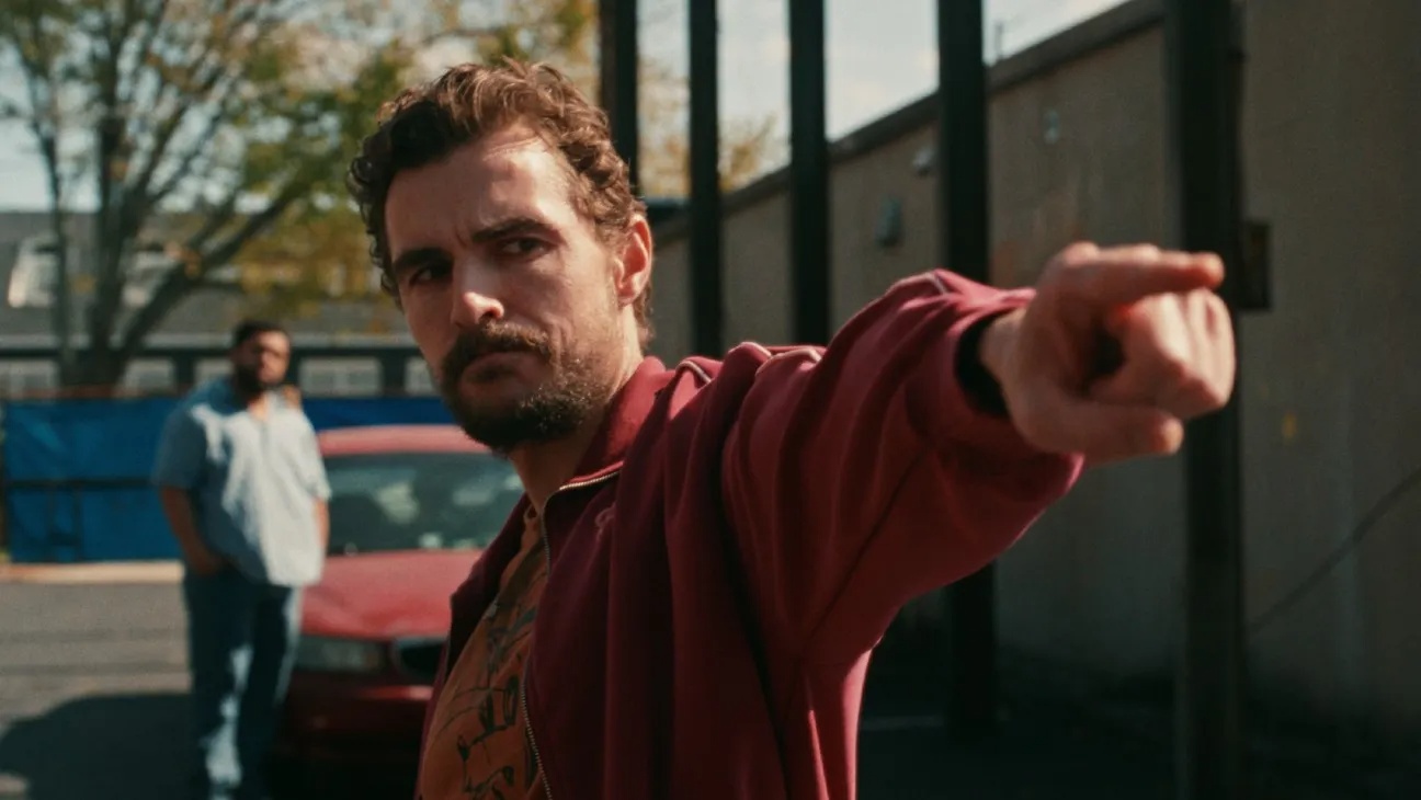 Man in a burgundy jacket throws a punch toward the camera in a parking lot, with a second man and a red car in the background.