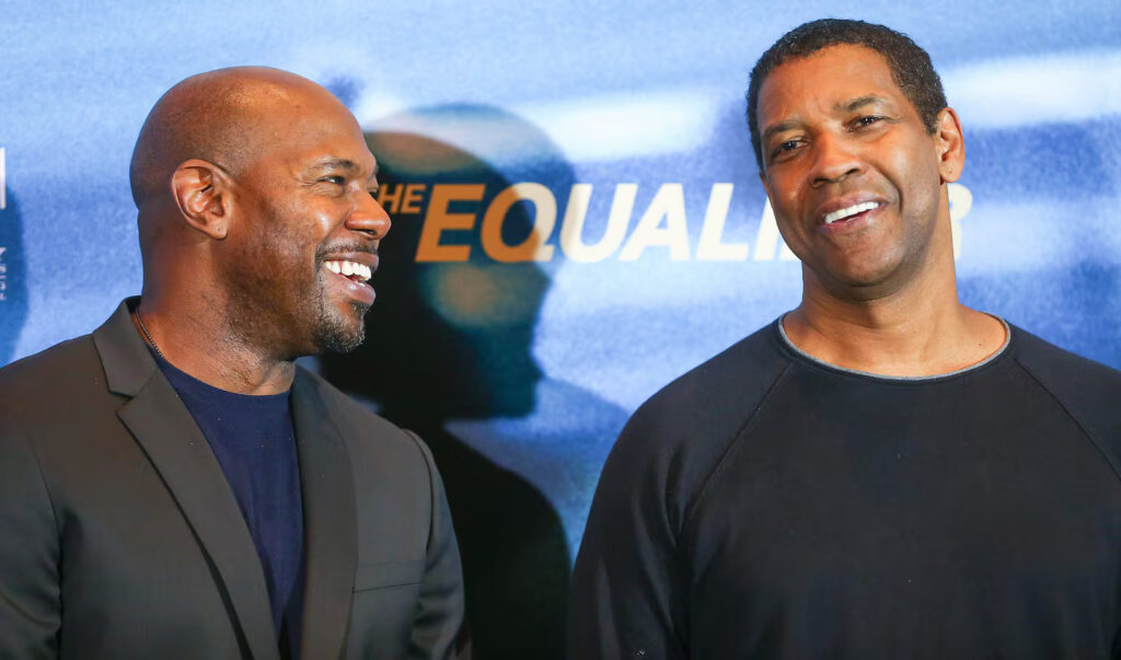 Two men smiling and chatting in front of a blue backdrop with the word 'EQUAL' visible, one in a dark blazer and navy shirt, the other in a black sweater.