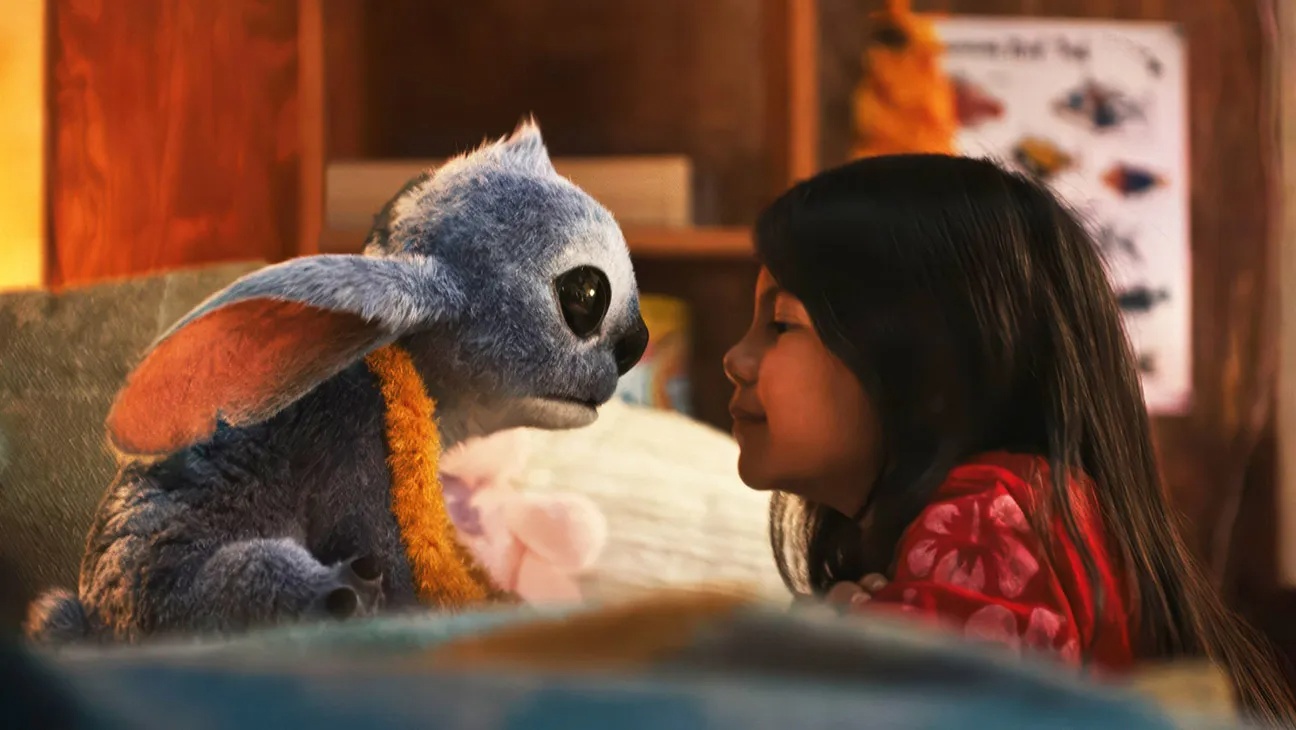 A young girl leans in to touch noses with a blue plush Grogu toy in a cozy indoor setting.
