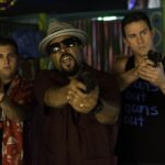 Three men aim handguns at the camera in a dim, colorful indoor setting; center man wears a hat and sunglasses and leads the pose.