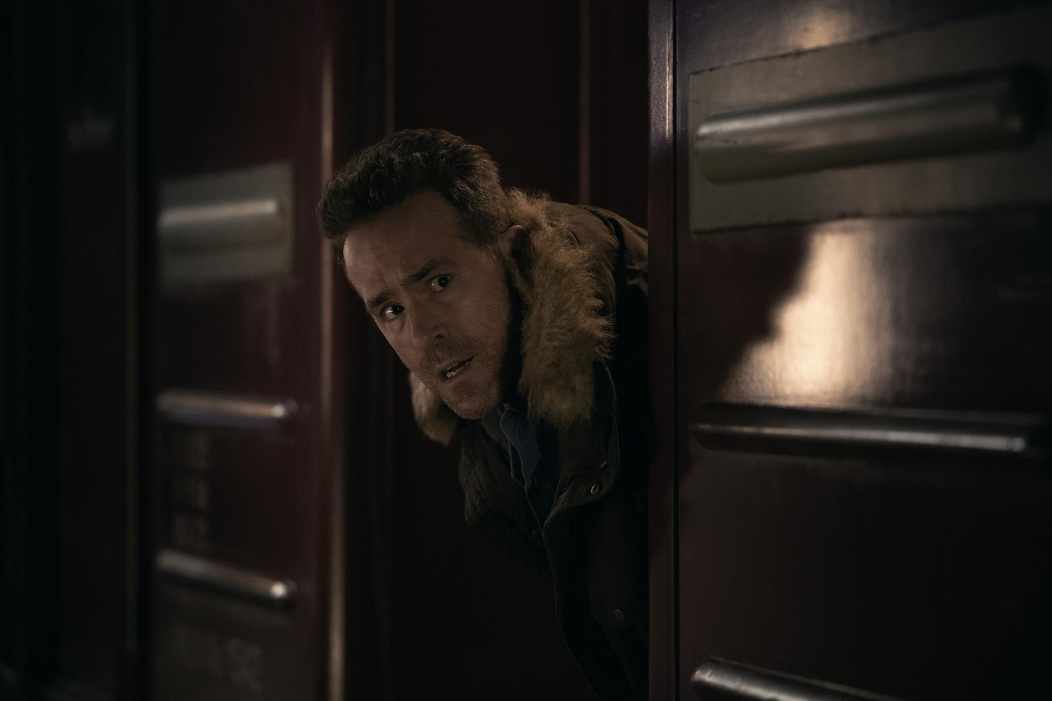 Man with a fur-clined jacket peeks from a doorway in a dimly lit, moody scene.