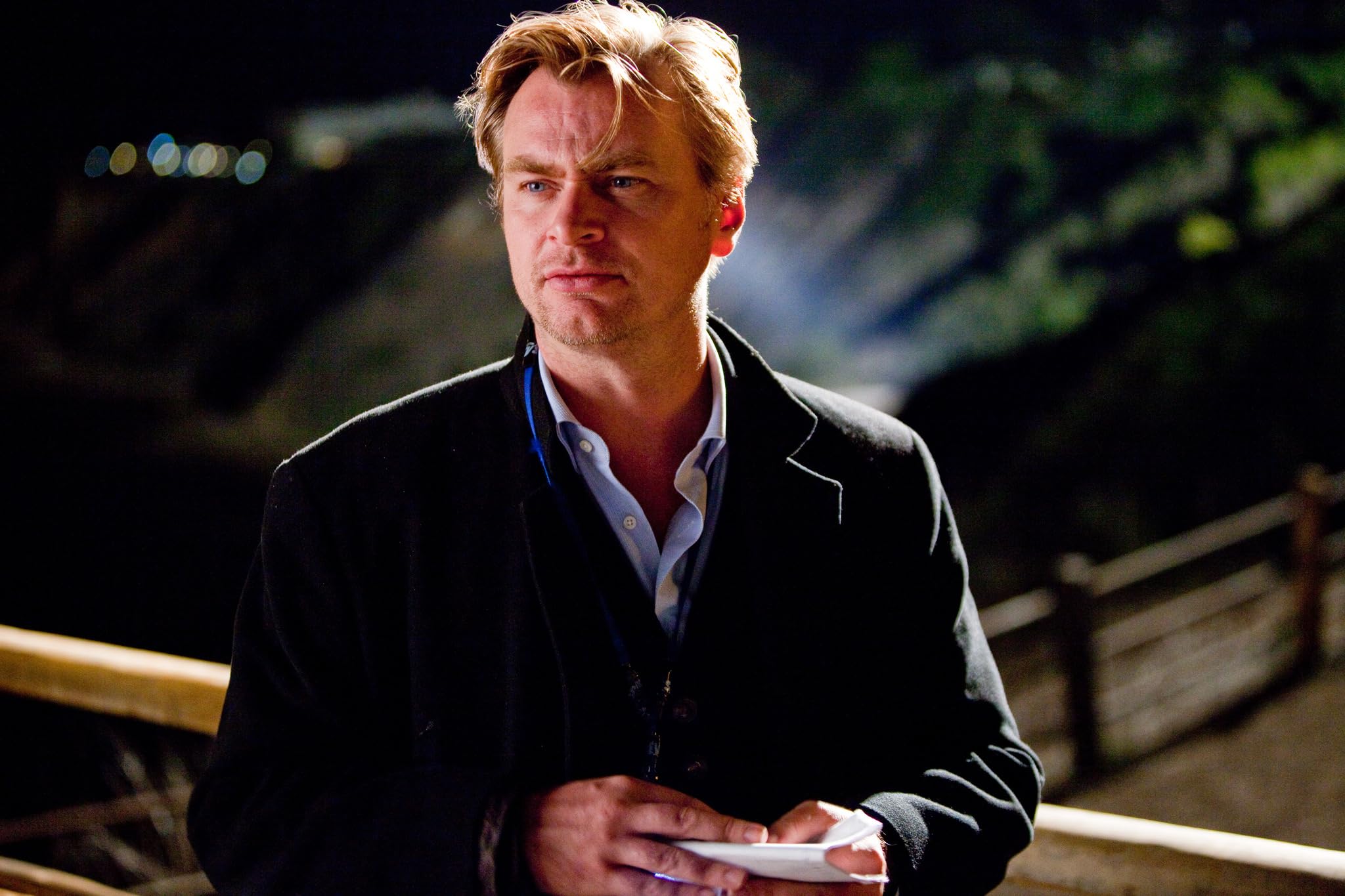 Man in a dark blazer holding papers outdoors at night, looking off to the side.
