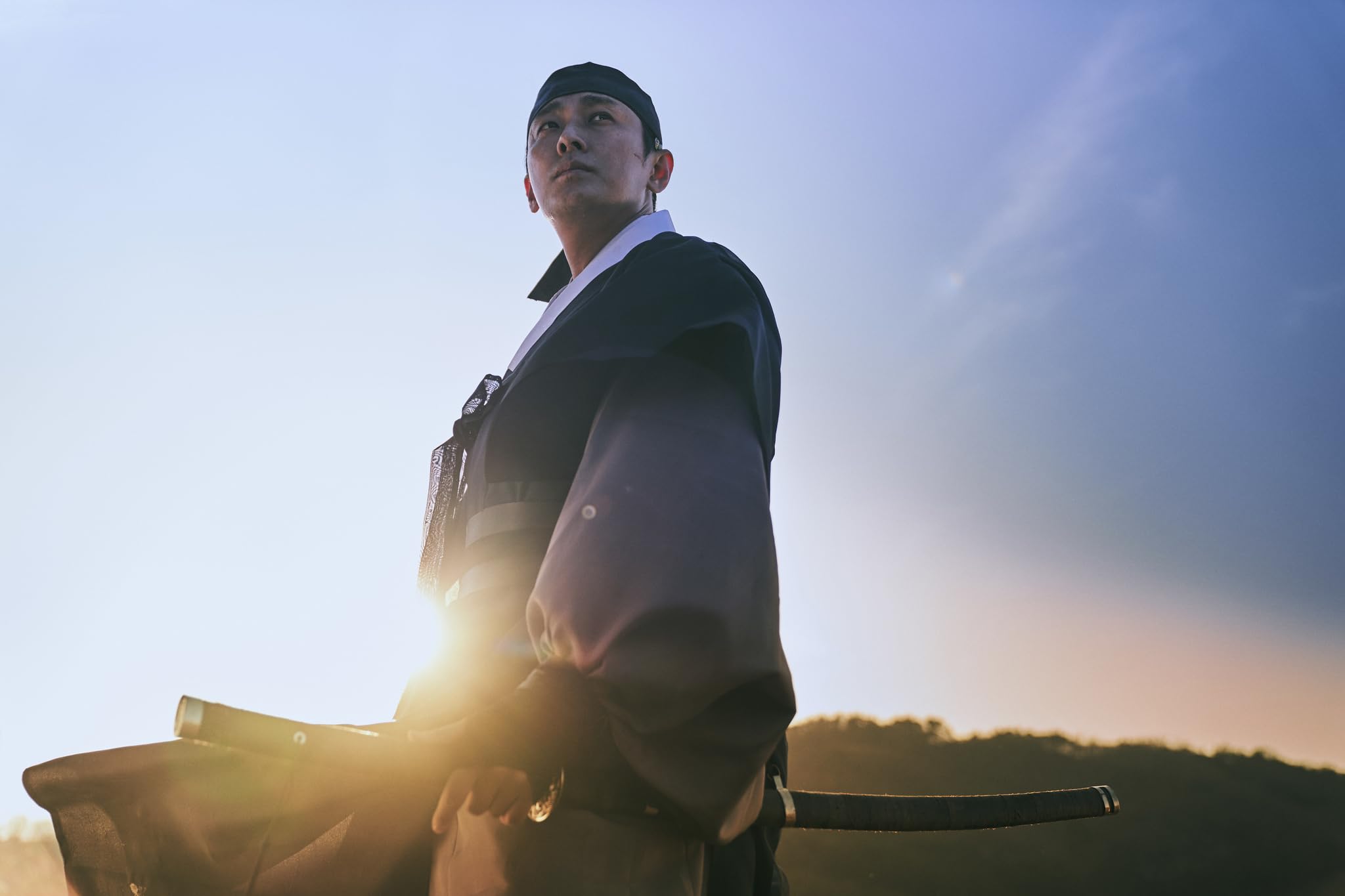 Worker in dark coveralls and cap standing outdoors, gripping a metal pipe with sun glare behind him.