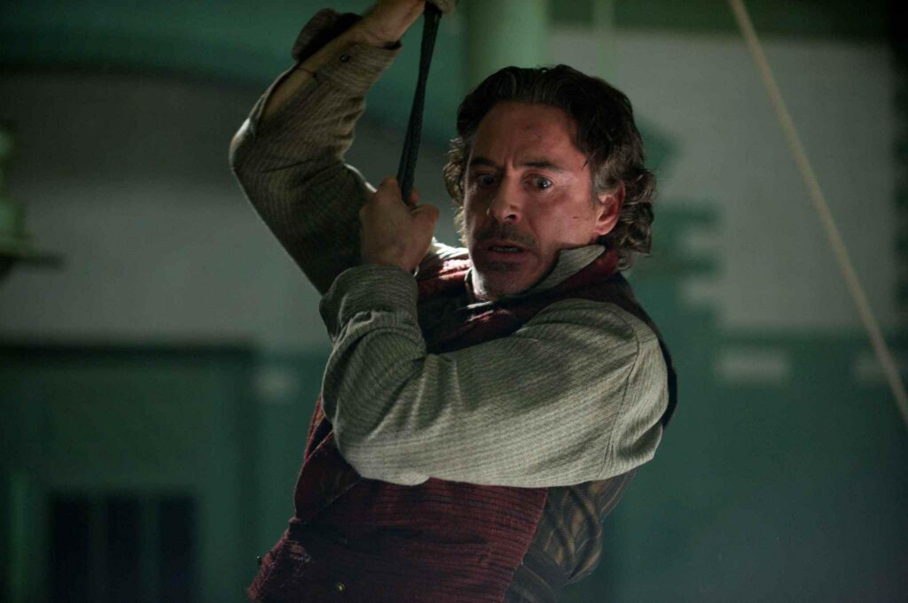Man with dark hair and stubble, wearing a striped sweater, grips a rope overhead in a dim, green-lit room, looking alarmed.