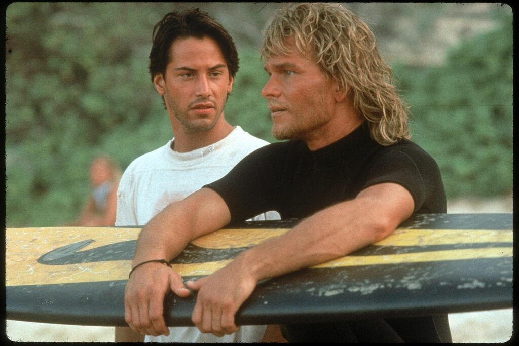 Two men leaning on a surfboard outdoors, one in a white shirt and the other in a black shirt, with a green, blurred background behind them.