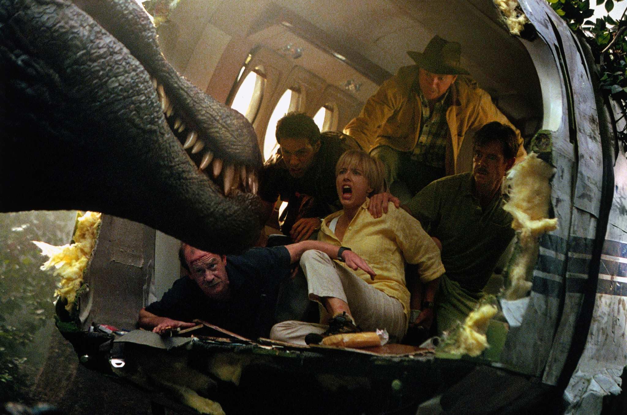 Group of people huddled inside a circular tunnel, terrified as a giant dinosaur-like mouth fills the frame.