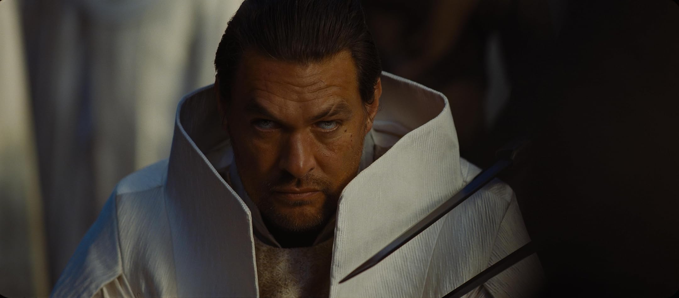 Man in a white high-collared robe holding a sword across his chest, with a stern, downward gaze against a dark background.