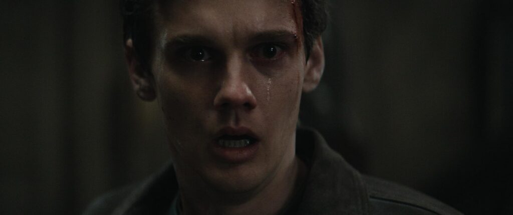 Close-up of a young man in a dark, tense environment, with a bloody cut above the eyebrow and a desperate, stunned expression.