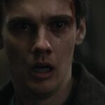 Close-up of a young man in a dark, tense environment, with a bloody cut above the eyebrow and a desperate, stunned expression.