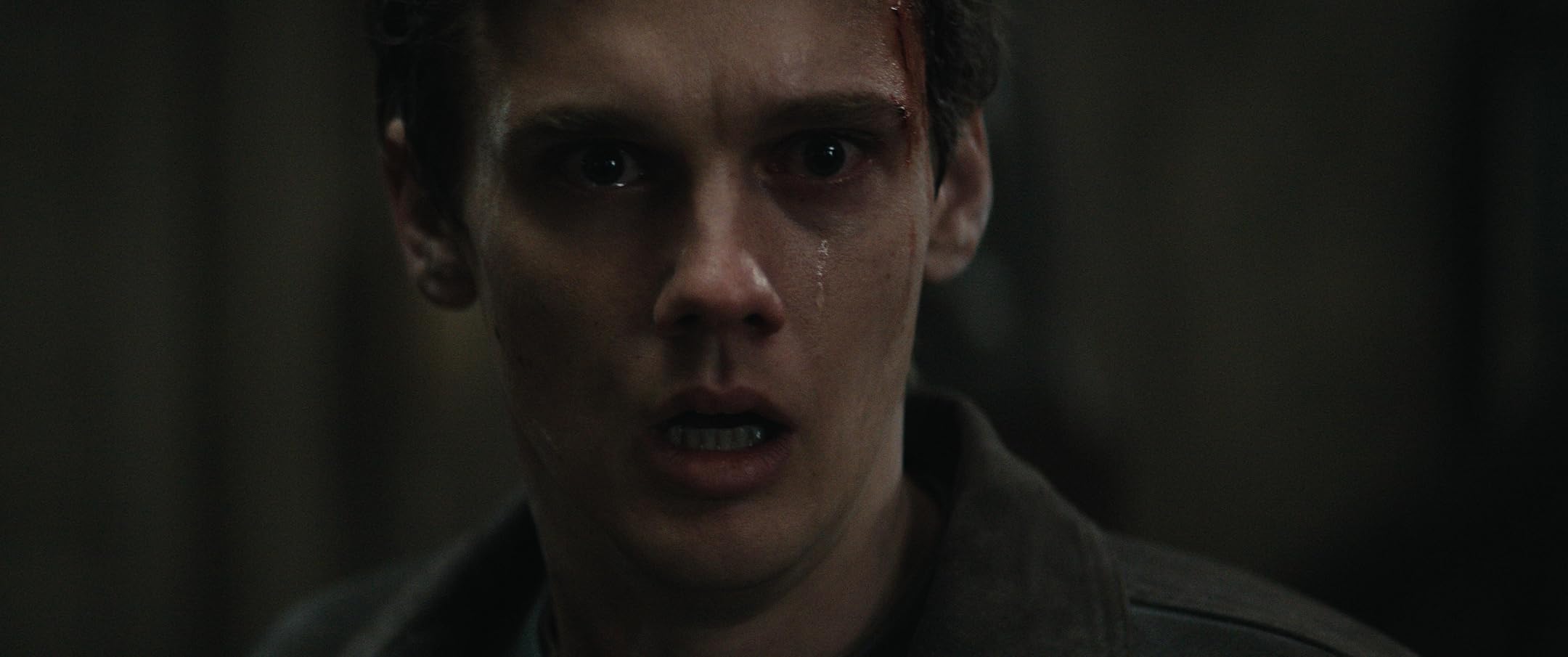Close-up of a young man in a dark, tense environment, with a bloody cut above the eyebrow and a desperate, stunned expression.