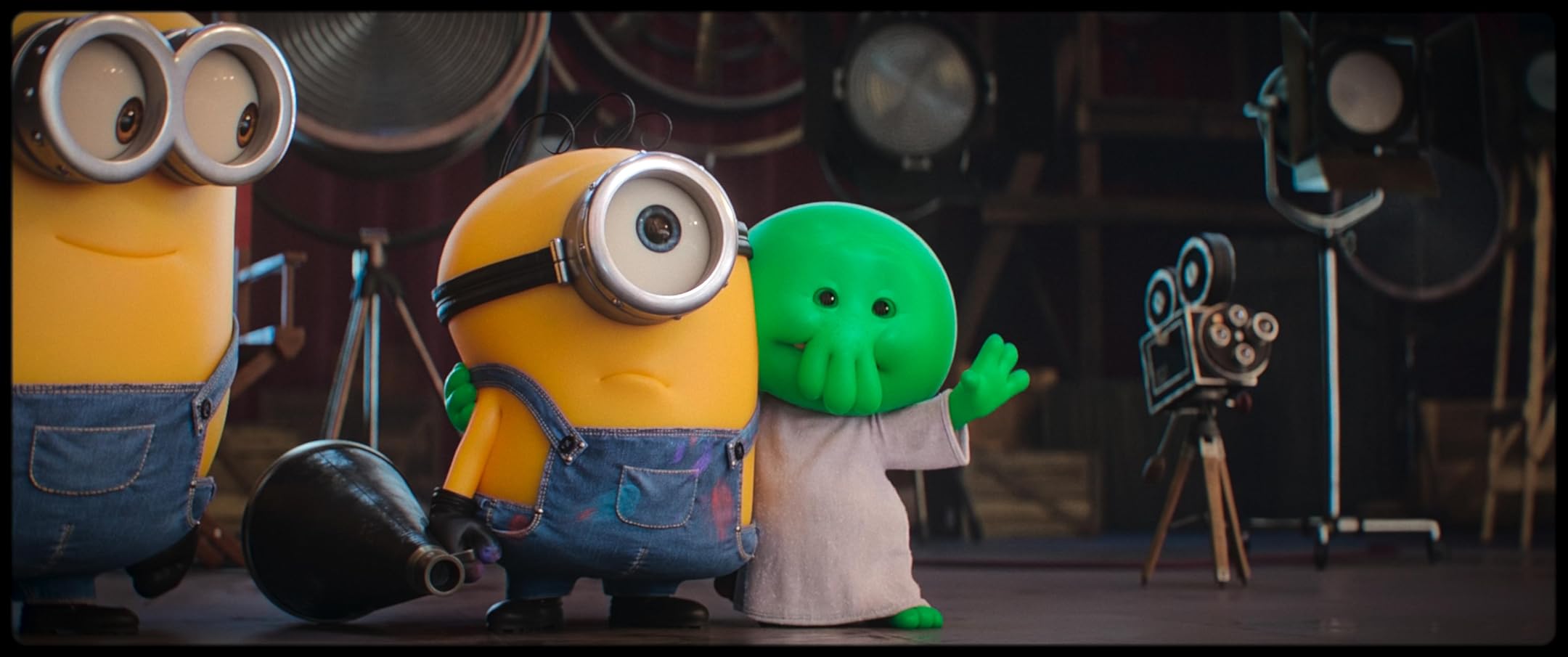 Two Minions stand on a film set with camera gear in the background; the yellow minion with goggles is hugged by a green alien-like character wearing a white shirt, both looking toward the camera.