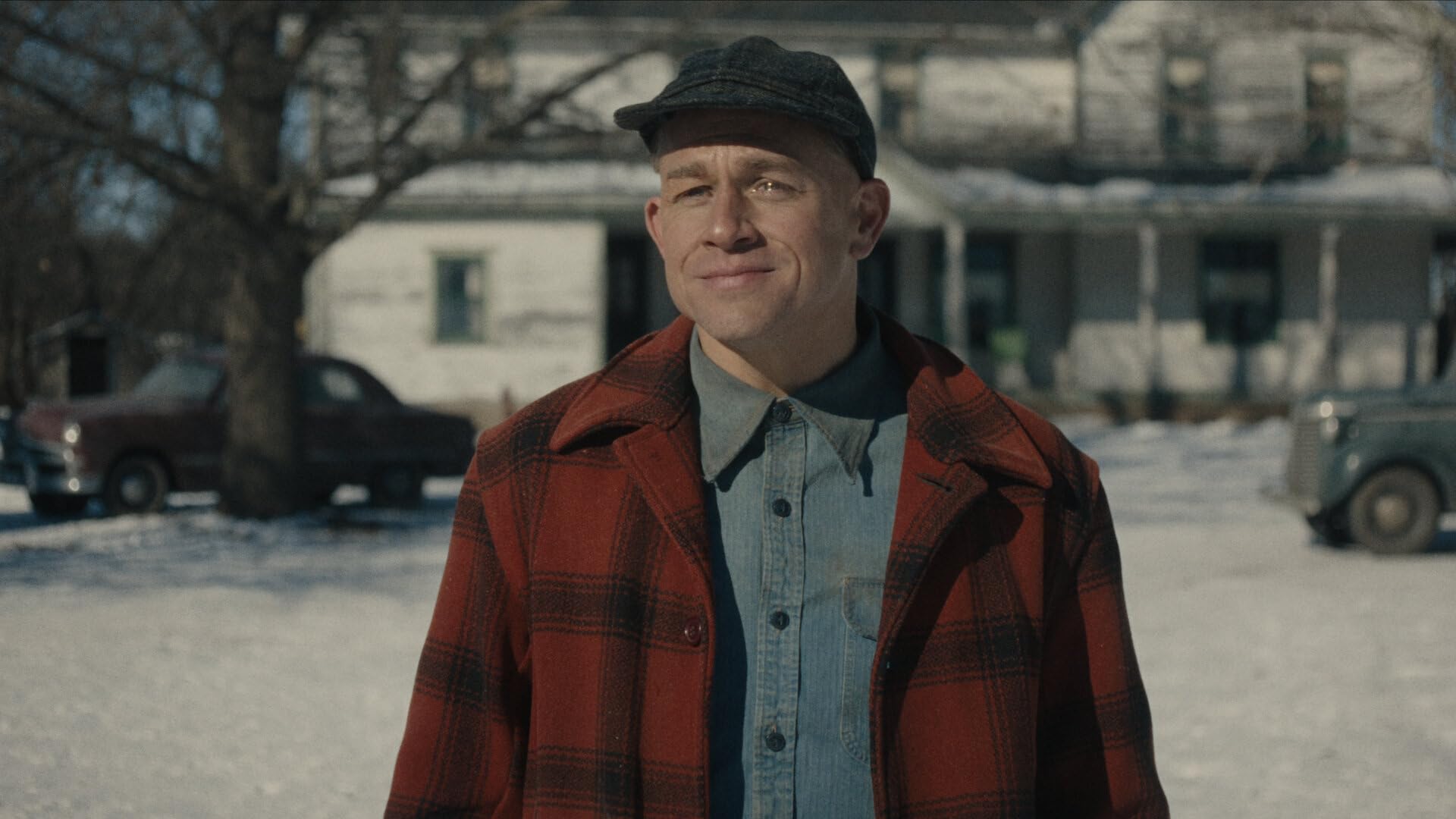 Man in a red plaid coat smiles outdoors in a snowy yard with a vintage house and cars in the background.